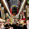 A busy covered shopping arcade beneath a large Kuromon lantern at Kuromon Ichiba Market in Osaka.