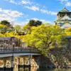 Osaka Castle rising above stone walls, a bridge and surrounding green trees.