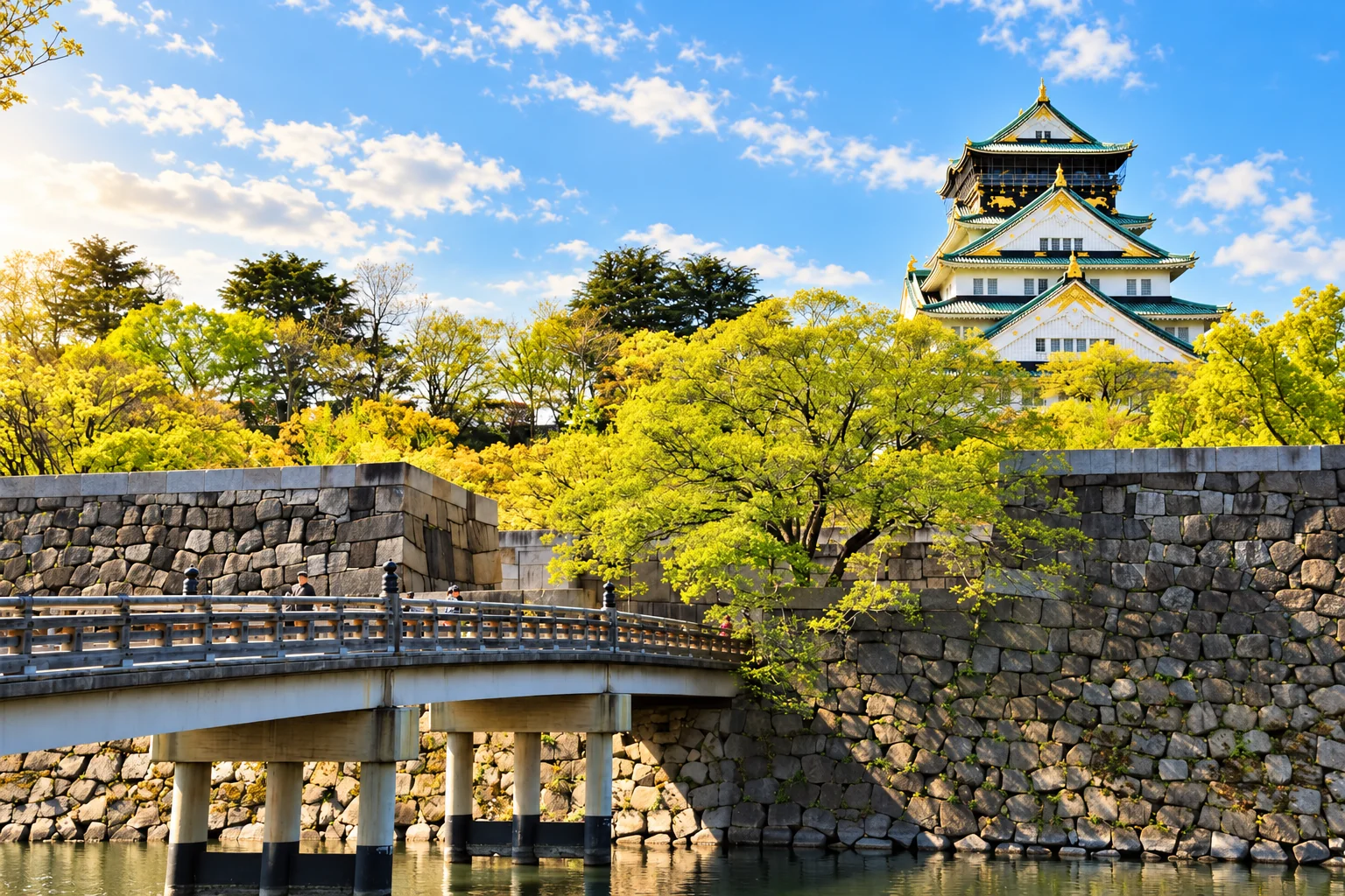 Osaka Castle rising above stone walls, a bridge and surrounding green trees.